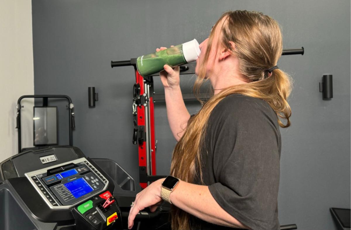 A woman drinking Cymbiotika plant protein from a shaker bottle while walking on a treadmill